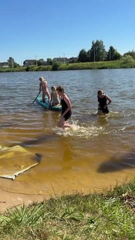 Three young females are in shallow, murky water near a sandy bank. One female in the foreground, wearing a black dress, is walking out of the water and splashing. Behind her, another female in a black dress is walking towards the shore. Further out in the water, two other females are on a paddleboard. The paddleboard is blue and has some design on it. One of the females on the paddleboard is holding a paddle. The environment appears to be a lake or river with a clear blue sky and trees and houses in the background. A dog with brown fur is visible in the foreground, facing away from the camera.