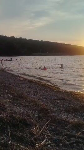 This media file captures a serene scene at a beach in Ustia, Moldova, likely during the late afternoon or early evening. The sky is a soft gradient of blue and grey, with hints of white clouds, suggesting mild weather conditions. The sun, not directly visible, casts a warm glow, illuminating the water and the treeline in the distance.

The foreground is dominated by a pebbly beach strewn with driftwood and dried vegetation, giving it a natural, somewhat untamed appearance. The water is calm and inviting, with gentle ripples reflecting the muted light.

Several people are enjoying the water. Some are swimming or wading, while others are seen with inflatable rings, indicating recreational activities. A group of individuals are clustered together in the water, likely socializing or playing. On the shore, a few figures are visible near the water's edge, and further back, a line of cars suggests a parking area for visitors.

The overall atmosphere is peaceful and relaxed, evoking a sense of leisure and enjoyment. The natural setting, combined with the presence of people engaging in simple pleasures, creates a tranquil beach experience. The lack of any visible text or prominent man-made structures, apart from the cars, emphasizes the natural beauty of the location.