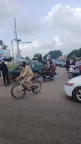 The video shows a busy intersection with several vehicles and people. A man on a bicycle is in the foreground. In the middle ground, a motorcycle with three people is seen passing by, followed by a green car and other vehicles. In the background, there are billboards with images of politicians and text that appears to be campaign material. A woman in a white shirt and black pants is holding a cell phone and looking towards the right side of the frame. Another woman is holding a large banner with campaign slogans and a picture of a politician. The sky is overcast with clouds. The scene appears to be a public street, possibly during a political event or rally, given the presence of campaign materials and the gathering of people.
