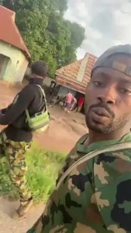 The video shows a man in camouflage clothing taking a selfie video, in what appears to be a rural area.  He is wearing a camouflage baseball cap and a light-colored shoulder strap. Behind him, another person in camouflage pants and a black shirt walks away.  In the background, there are buildings, and a group of people near one building. There is also some green vegetation. The sky is overcast. There are audible sounds that appear to be shouting or gunfire.