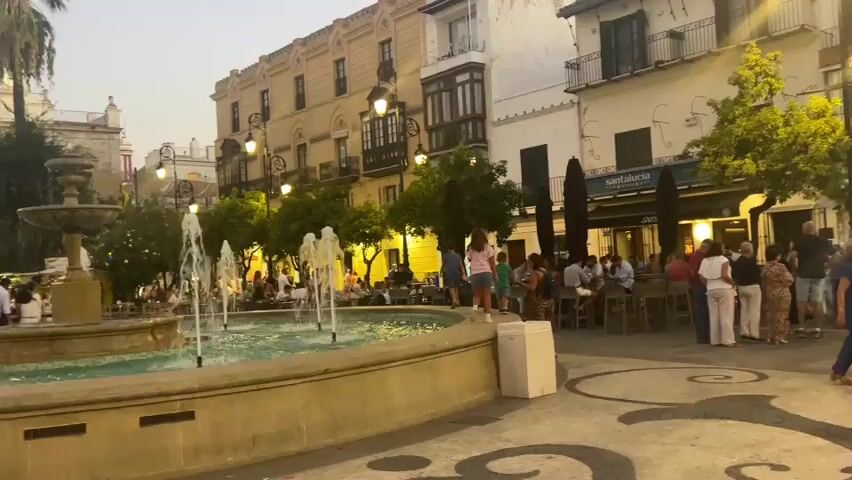 This video shows a fountain in a city square with people gathered around it. The fountain has multiple jets of water spraying upwards. In the background, there are buildings with balconies and awnings, as well as trees. Several people are seated at tables outside a cafe, and others are standing and walking around the square. The lighting suggests it is dusk or evening.