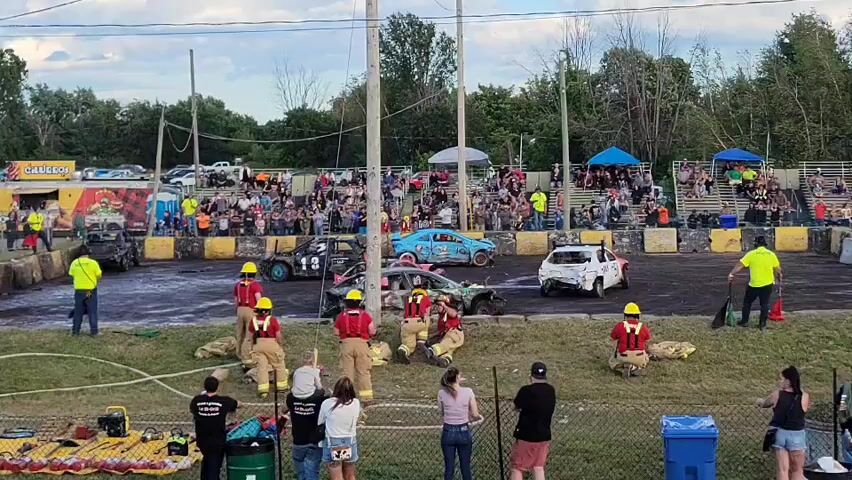 This is an aerial shot of a demolition derby event. There are several wrecked cars in an arena. A crowd of spectators watches from bleachers. A group of people in firefighting gear are on the ground near the arena, holding hoses. Other people are standing and watching the event. The environment appears to be an outdoor event space, possibly a fairground, with trees and a fence visible in the background. The sky is overcast.