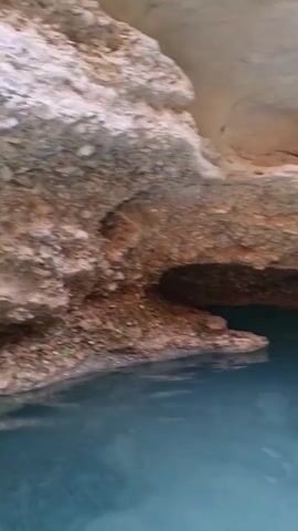 The video captures an underwater view looking towards a rocky overhang. The water is clear and blue, revealing a pebble-strewn bottom. The rock formations above are textured and varied in color, ranging from light beige to reddish-brown. The camera appears to be moving slightly, creating subtle ripples on the water's surface. There are no people or other objects visible in the frame.