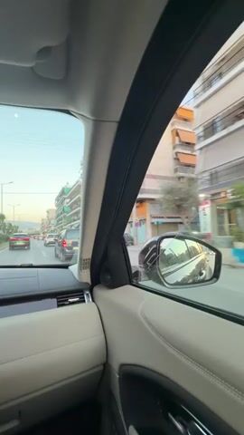 A video shows a woman with long brown hair, wearing glasses and a green jacket, sitting in a car. She is smiling and looking at the camera. At one point, she blows a kiss to the camera. Views from inside the car alternate with quick shots out of the side window, showing urban buildings with balconies and other cars on a street. The car appears to be moving, as the external scenery is blurred. A man's arm and shoulder, wearing a white t-shirt, are briefly visible in the background, suggesting he is also in the car. The time of day appears to be daytime, with natural light illuminating the scene. The setting is an urban environment, specifically the Municipal Unit of Tavros, Greece. The woman appears happy and playful.