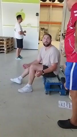 A man with a beard and a light-colored t-shirt and dark shorts is sitting on a blue pallet. He is looking towards the camera with a neutral expression. His left leg is crossed over his right leg. He is wearing white sneakers and white socks. Another man in a red t-shirt and blue shorts is standing to the right of the frame, facing away from the camera. He has his hands on his hips. In the background, another man in a white t-shirt and black shorts is walking away from the camera. The environment appears to be a warehouse or storage facility, with pallets and shelves visible. There is a sign with a green apple logo and text that reads "Fructe & legume" on the wall.