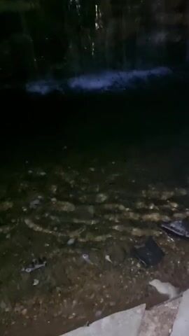 A body of water is seen at dusk. The water is dark and appears to be murky. There are rocks and debris visible at the bottom of the water. In the background, there is a waterfall cascading down a rocky cliff face. The water from the waterfall is illuminated by artificial lighting, giving it a blueish glow. Trees and a light fixture can be seen at the top of the cliff. The overall scene is dimly lit and appears to be a man-made feature in a natural setting.