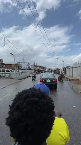 The video shows a person riding on the back of a motorcycle on a wet road in an urban environment. The rider is wearing a yellow shirt and a blue baseball cap, and the person recording is behind them, capturing the back of their head. The road has light traffic, including a black car, other motorcycles, and a yellow tricycle. The sides of the road are lined with buildings, utility poles, and streetlights. The sky is blue with scattered white clouds. The video appears to have been taken during the daytime.