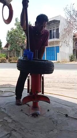 A man is shown operating a tire changer machine, which is a red, mechanical device. The man is wearing a maroon t-shirt and dark pants. He is using a lever attached to the machine to work on a tire that is mounted on the machine. The tire is black and appears to be a car tire. The scene is outdoors, in what appears to be a workshop or service area, with a paved ground surface. In the background, there is a road with some vehicles, including a white car and a white truck. There are also buildings and trees visible in the background. The overall impression is that of a tire repair or changing operation.