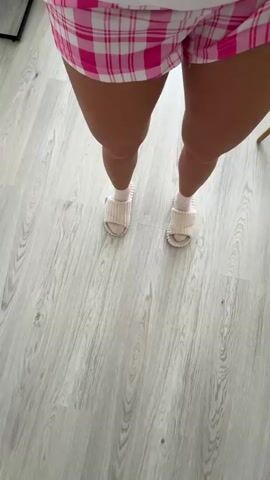 A first-person perspective video shows a person's legs and feet walking on a light-colored wooden floor. The person is wearing pink and white plaid shorts and white fuzzy slides over white socks. The camera angle is tilted downwards, focusing on the lower body and the floor. In the lower left corner of the frame, a pair of black shoes is visible on the floor. The scene appears to be indoors, possibly a home environment.