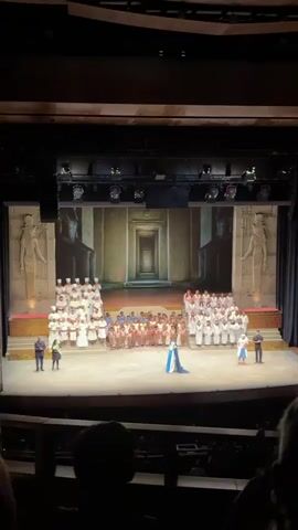 The video shows a wide shot of a theatrical stage from the perspective of the audience in a darkened theater. The stage is set with an Egyptian theme, featuring painted backdrops depicting pharaoh statues and a long hallway. In the center of the stage, a large group of performers is arranged in rows. They are wearing a variety of costumes, including white robes, brown tunics, and a blue and black gown.  Several individuals in business attire stand at the edges of the stage. The audience is visible in the foreground, with heads and shoulders silhouetted against the light from the stage. After an undetermined event, the audience applauds.