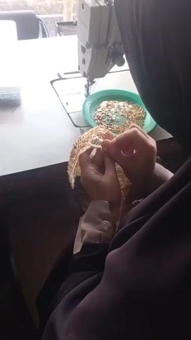 A person wearing a dark hijab and brown clothing is sitting at a table, sewing. They are holding a piece of gold-colored fabric with intricate patterns in their hands and using a needle and thread to attach small, gold-colored embellishments. To the right of the fabric is a green plastic plate filled with these embellishments. A white sewing machine is visible behind the plate on the table. The table appears to be a light color. The background is blurry but suggests an indoor setting with some light coming from the left.