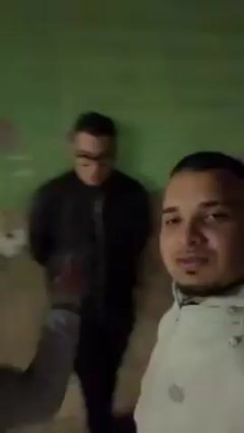 The video shows a man filming himself with a group of people in the background. The man is wearing a grey zip-up jacket. He has short dark hair and a goatee. The other people are sitting on steps. A woman in a pink jacket is on the left. Next to her is a man in a white sweatshirt. To the right of the man is a woman in a green shirt. The background appears to be an alleyway with a green wall with graffiti in the beginning of the video. The man speaks in the video and makes a gesture with his finger towards the camera.