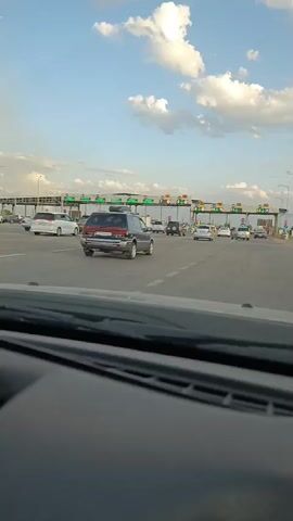 The video shows a view from the dashboard of a car driving towards a toll booth plaza. There are several lanes of traffic with cars in them. The toll booths are overhead structures with lights and signs. The sky is blue with white clouds. The traffic appears to be moving slowly.