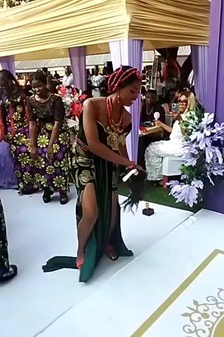 At an outdoor event venue, a woman in a black and green strapless dress with a high slit and red beaded jewelry dances on a white platform, holding a dark accessory. Other women dressed in matching floral print dresses with black lace details stand nearby. The event space is decorated with a beige awning, purple fabric, floral centerpieces, and sparklers placed along the platform edge. People are seated at tables in the background, observing the performance.