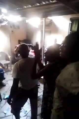 The video shows a group of people gathered indoors, in what appears to be a casual setting. The interior looks somewhat run-down, with an exposed ceiling structure. People are standing, sitting, and moving around. Some individuals are more visible than others due to the angle and lighting. There is a small table with items on it, possibly refreshments. The atmosphere seems relaxed and social, though specific interactions are hard to discern due to the video quality and camera movement.