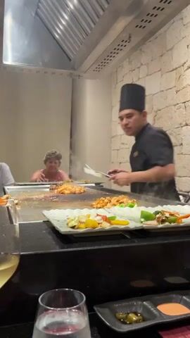 A chef in a black uniform and chef's hat is cooking food on a large, flat griddle. The griddle is in front of a wall made of light-colored stone. Two people, a woman with short, gray hair and a man with a white shirt, are sitting at the griddle, watching the chef. The chef is using two spatulas to move food around on the griddle. In the foreground, there are plates of prepared vegetables, glasses of white wine and water, and small dishes of condiments. Steam rises from the griddle. The overall environment appears to be a restaurant with a hibachi-style grill.