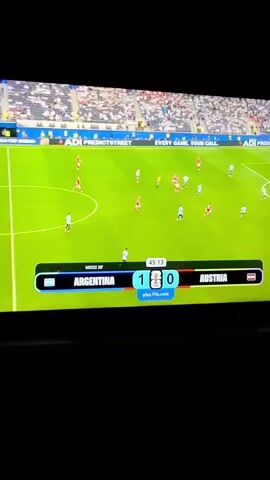 This media file captures a screen displaying a soccer match between Argentina and Austria. The score is 1-0 in favor of Argentina. The game is in its 45th minute and 18th second. The field is green, with players in red and white uniforms scattered across it. The stadium is filled with spectators, though their faces are not discernible. In the foreground, a digital scoreboard shows the team names, scores, and a countdown timer. Visible text includes "ADI PREDICTSTREET" on advertising boards and "play.fifa.com" below the score. The overall scene depicts a competitive sporting event. There are no visible cues to indicate the time of day or weather conditions. The location is identified as Büyükçekmece, Turkey, but this context is external to the visual information in the media file itself.