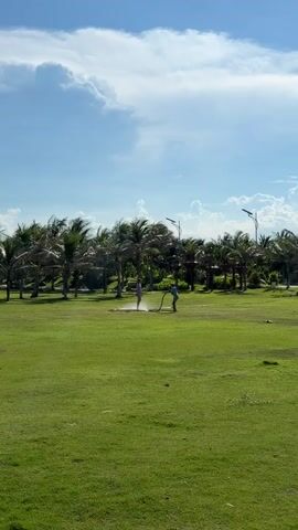 Two individuals are visible in a grassy field with palm trees in the background under a partly cloudy sky. One individual, wearing a pink top and white bottoms, stands near the center of the frame. The other individual, wearing a blue shirt and black shorts, is to the right and appears to be holding a bicycle. This individual is also spraying water from a hose, creating a visible spray arc in the air. The overall scene suggests an outdoor recreational area or park.