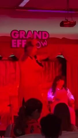 A performance is taking place on a stage. There is a banner at the top of the stage that reads "GRAND EFE SHOW." The stage is lit with red and blue lights. On the stage, a person in a long, light-colored garment is performing, holding a microphone. To the left, another person in a black top and light-colored pants is visible. To the right, a person in a light-colored top and dark pants is also visible, and another person in a darker outfit. In front of the stage, an audience is seated, with their backs to the camera. The lighting changes between red and blue throughout the short clip.