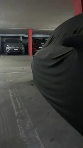 A camera pans across a dark car cover in a parking garage. The camera then moves closer to reveal the front of a gray car underneath the cover. The car's hood is partially open. The parking garage appears to be underground or enclosed, with concrete walls and red-painted support columns visible. Other cars are parked in the background.