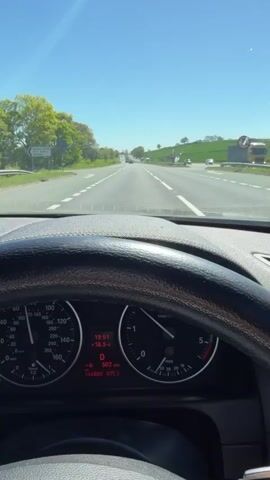The video depicts a driver's perspective while driving on a highway. The dashboard, including the speedometer and other gauges, are visible. The speedometer reads around 50 mph. Through the windshield, we see a straight, multi-lane highway with light traffic, a blue sky, and green fields bordering the road. The weather is sunny and clear.
