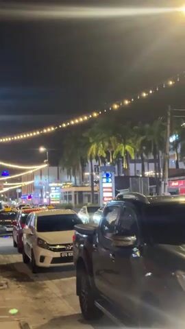 A vertical, nighttime, low-light video shows a street scene with a line of cars. The camera is positioned at an angle, looking down the street from sidewalk level, with a black pickup truck in the foreground on the right. String lights are suspended across the street, illuminated by a warm glow. Palm trees line the sidewalk on the left side of the street. Buildings with various signs and lights are visible in the background, including what appears to be a gas station. Several cars are parked or moving slowly in traffic, including white sedans, red cars, and a yellow taxi. The overall atmosphere is that of a busy street at night, possibly in a commercial or entertainment district.