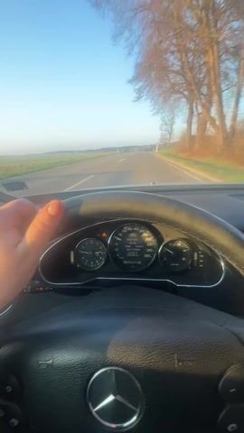 The video shows a driver’s POV of a car ride on a rural road. The car's dashboard is visible, with a speedometer that goes up to 260 km/h. The steering wheel has a prominent Mercedes-Benz logo. Outside, the road is straight, flanked by fields and trees, with a clear blue sky above. A warning sign is also visible on the right side of the road. Electronic music plays in the background.