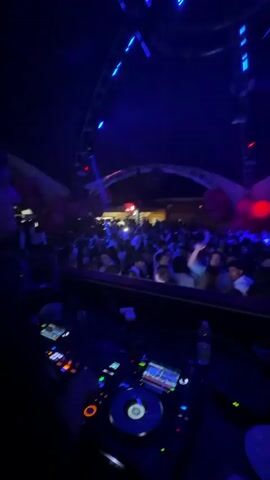 The video shows a DJ performing in front of a large crowd at a nightclub. The DJ is wearing a black shirt and is operating a Pioneer DJ mixer and turntables. The crowd is dancing and cheering, with many people holding up their hands in the air. The club is dark, with blue and white lights flashing, and some firework machines in front of the DJ. The camera is positioned behind the DJ, providing a view of the crowd and the DJ's hands as they manipulate the equipment. There is a bottle of water on the counter in front of the DJ gear.