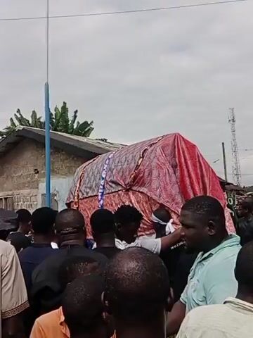 A funeral procession is underway, with a group of people gathered around a large, ornate coffin covered in red and gold fabric. The coffin is being carried by pallbearers, and the crowd appears to be observing with solemnity. The scene is set outdoors, with a cloudy sky and some trees visible in the background. The people in the crowd are dressed in a variety of clothing, with some wearing traditional funeral attire. The overall atmosphere is somber and respectful.