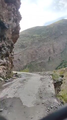 A first-person perspective video shows a dirt road winding through a mountainous landscape. The road is uneven and rocky, with loose gravel and dirt. To the left, a steep, rocky cliff face rises up. To the right, a steep drop-off into a valley is visible, with a further mountain range in the background. The sky is overcast with clouds. The video captures the bumpy motion of traveling on this road.