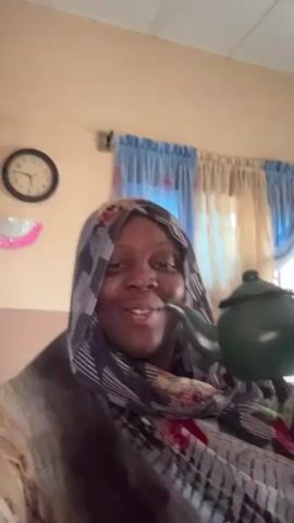 A woman is in the frame from the chest up, smiling and holding a small green teapot up to her mouth. She appears to be pretending to drink from the teapot, holding it with her right hand. She is wearing a headscarf with a pattern of black, white, and gray stripes, and a floral pattern. The background includes a wall with a beige-colored top portion, a clock reading 10:10, and a window with blue and beige curtains. A small pink decoration is visible on the wall to the left of the clock.