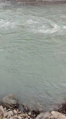 This video shows a choppy river with some white water rapids near the top of the frame. The riverbed is rocky and covered in stones and some debris on the right side of the frame. The water appears murky and grey.