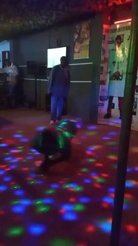 The video shows a dimly lit room with a dance floor illuminated by colored lights. In the foreground, a person is on all fours, seemingly dancing or performing a breakdance move. In the background, two other individuals are visible. One person, wearing a blue sweatshirt and pants, stands near a television screen. Another person, dressed in a white outfit, stands to the right of the frame, possibly observing or participating. Posters are visible on the wall behind them. The atmosphere appears to be that of a nightclub or a party.