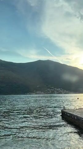 The video shows a panoramic view of a body of water, with mountains in the background. The water is a deep blue, and the mountains are covered in lush green vegetation. The sky is a clear blue, with a few white clouds scattered across it. In the foreground, there is a concrete pier with a few cars parked on it. One of the cars is a black Fiat Panda covered with a white tarp. Another car is a white Mazda CX-5 with a license plate visible. The video appears to have been filmed at sunset, as the sun is setting behind the mountains, casting a warm glow over the scene. There is a boat docked at the pier. A plastic water bottle is lying on the pier.