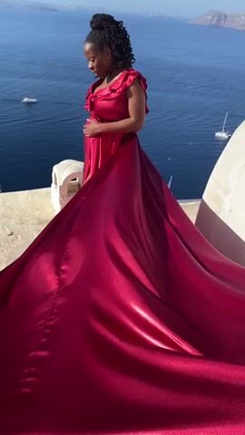 The video shows a pregnant woman in a long, flowing red dress standing on a balcony overlooking the ocean. The woman is of African descent and has her hair pulled up in a ponytail. She is holding her belly and smiling at the camera. There are boats in the water and islands in the background. The scene appears to be in a sunny, warm climate.