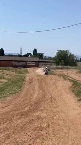 The video shows a dirt bike rider in full gear, including a white helmet and black and white riding suit, riding a white and black motorcycle on a dirt track on a sunny day. The rider kicks up dust as they approach the camera on a small hill. Behind the rider is a single-story building with a brown roof and a sign. After the rider passes, the camera pans right to show more of the dirt track with other riders and an "AHAMAY" flag, with a city visible in the distance.