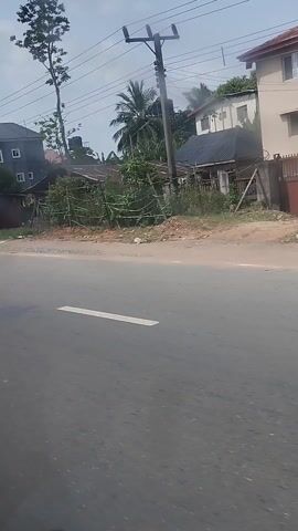 The video shows a roadside scene with various buildings and structures. The camera appears to be moving, capturing the scene from a vehicle traveling along the road. Several buildings are visible, including residential houses of varying sizes and designs, a filling station, and small roadside shops. There are also trees and power lines along the roadside. People are seen walking along the side of the road. The overall atmosphere appears to be daytime in a residential or commercial area.