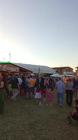 This video shows an outdoor gathering at dusk, likely an evening festival or fair in Arbia, Italy. A large crowd of people are milling around on a grassy field. In the background, several tents and structures are set up, including one labeled "Birreria" with images of beer mugs. To the right, there's a large inflatable water slide, suggesting it's a family-friendly event. People of all ages are present, some standing in lines at the food stalls, others conversing, and children are seen playing. The sky is a clear, pale blue, transitioning to a warm glow near the horizon, indicating the time is around sunset. The overall atmosphere appears relaxed and festive, with people enjoying the evening outdoors.