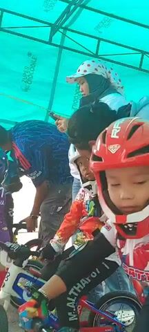 A group of children and adults are gathered outdoors under a green tarp. Several children are wearing helmets and cycling gear, suggesting they are participating in or preparing for a cycling event. One child in the foreground, wearing a red helmet, is looking directly at the camera. Another child in a white helmet is visible behind the first child. Adults are standing or leaning in the background, some appearing to be supervising or cheering. The overall atmosphere seems to be one of excitement and preparation for an activity.