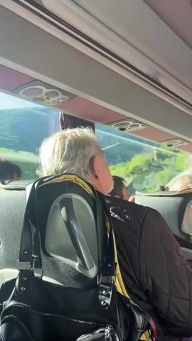 From the perspective of a passenger on a bus, the video shows the backs of other passengers sitting in front. Many are older adults with gray or white hair. The bus interior is gray with black seats outlined in yellow. A black purse hangs on the back of the seat in the foreground. The bus moves through a bright, green environment as seen through the windows, which are lined with a light pink curtain. The interior lights dim, creating a darker atmosphere.