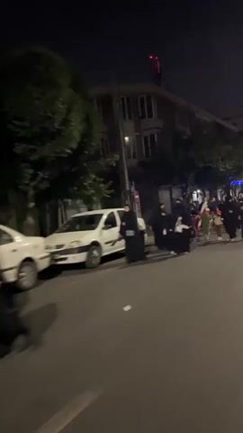 This video captures a nighttime scene on a city street. A group of people are walking along the road. Several cars are parked on the side of the street. Trees line the sidewalk. The lighting suggests it is dark outside, with artificial lights from streetlamps and buildings illuminating the area. Some individuals in the group are holding red flags. The people are dressed in dark clothing, with some women wearing hijabs. The overall atmosphere appears to be that of a procession or march.