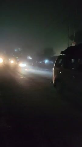 The media file depicts a dimly lit, foggy night scene on a street in Kulang, India. The primary subject is a white car driving through the fog, its headlights cutting through the haze. The car appears to be a hatchback, possibly a Maruti Suzuki Alto, given its shape and commonality in India.

The setting is a road with a paved surface that appears wet or damp, reflecting the car's headlights. On the right side of the road, there are buildings, one of which has a sign with the number "192" prominently displayed in red. This could be a house number, a shop number, or a street marker. Other structures are visible behind the car, obscured by the fog. Power lines are also visible overhead.

The atmosphere is thick with fog, significantly reducing visibility and creating a somewhat eerie or mysterious mood. The time of day is clearly night, as indicated by the darkness and the artificial lighting from the car and buildings. The weather is characterized by fog, which is dense enough to soften and diffuse light sources.

The activity is simple: a car is in motion on a street at night, navigating through the fog. No people are clearly identifiable as distinct subjects, though there may be occupants within the car, their presence suggested by the headlights and internal illumination. The overall impression is one of a solitary journey through challenging visibility conditions. The presence of the "192" sign provides a specific, albeit limited, location cue.