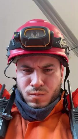 A man wearing a red helmet with a headlamp, an orange jumpsuit, and a gray neck gaiter is shown in a close-up, head-and-shoulders shot. Two Motorola radios are strapped to his chest. He makes a series of expressions, starting with a neutral look, then scrunching his face up in distaste, followed by a wide, slightly goofy smile. The background is a blurred, light gray ceiling or wall.