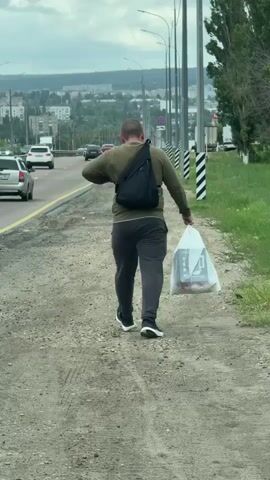 A man walks away from the camera on a dirt path next to a busy highway. He is wearing a green long-sleeved shirt, dark grey pants, and black and white athletic shoes. He has a black sling bag on his right shoulder and is carrying a clear plastic bag with a white object inside in his right hand. The highway has multiple lanes of traffic moving to the left. In the background, there is a city with buildings and trees. The sky is overcast.