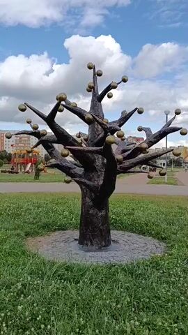 A statue of a stylized tree with golden spherical fruits sits in a grassy area. Behind the statue, a playground with slides and swings is visible, along with apartment buildings and a street. The sky is blue with fluffy white clouds. The camera slowly pans to the right.