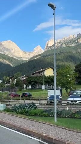The video shows a street scene in a mountainous area. In the foreground, the rear of a silver Volkswagen Touareg is visible, parked on the left side of the road. The road is paved with cobblestones. Beyond the road, there is a green lawn with some plants and flowers. A tall street lamp stands on the right side of the lawn. Behind the lawn, there are several houses, some with wooden balconies adorned with flower boxes. One house is painted yellow, and another is white with brown trim. In the background, a dense forest covers the lower slopes of majestic, rocky mountains, with clear blue sky above. Several cars are parked along the street. A large white signpost with directions to various towns and attractions is visible on the right side of the video. A person appears to be standing outside one of the houses. The lighting suggests it is daytime, likely late afternoon or early evening.