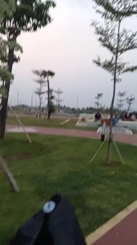 The video shows a group of people walking in a park near a body of water. The sky is overcast, and the lighting suggests it is either dawn or dusk. There are trees and grass throughout the park, with paved paths winding through the area. Some people are seated on benches, while others are walking along the paths. In the background, a line of people can be seen near the water's edge. The camera moves, indicating that the person filming is walking and recording their surroundings. Some individuals are wearing hijabs, suggesting a cultural context.