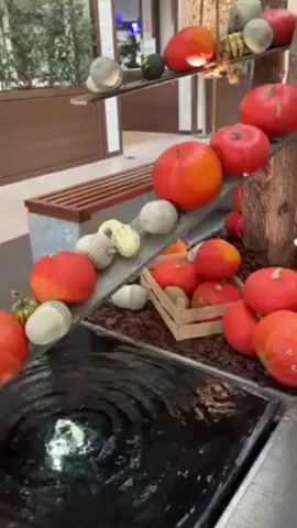 The camera provides a stable shot of an indoor decorative fountain arrangement. A square black basin holds shimmering water with a central fountain jet creating concentric ripples. The fountain sits among decorative elements including an angled ramp made of light gray wood, adorned with various types of pumpkins and gourds. Many pumpkins are round, with bright red-orange hues, while others are spherical and covered in a white mesh material. Some pumpkins are arranged in a small wooden crate. The ramp ascends to the right, supported by a simulated tree trunk and displaying pumpkins of varying sizes and colors. In the background, an indoor bench and glimpses of a shopping mall environment are visible.