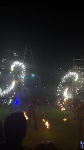 A wide shot at night shows a group of men performing a fire and light show in front of a crowd. On the left, a man spins glowing lights that create spiral patterns. In the center, a man manipulates multiple flaming objects, surrounded by several small fires on the grass. Another man on the right also performs with fire. In the background, a large screen displays abstract blue and white light patterns. The environment is dark, with the performers and their acts illuminated by the light and fire. The crowd's heads are visible in the foreground.