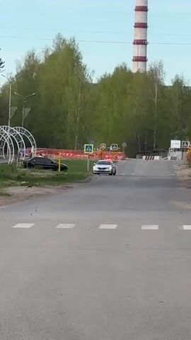 The video shows a street scene on a sunny day. In the background is a tall, industrial smokestack with red and white stripes.  A road with a pedestrian crossing runs across the foreground. A white car with flashing lights is stopped in the middle of the road, just before the crossing. A black car is parked on the grass to the left of the road, near a modern, illuminated white arch.  Two people are walking through the arch. Traffic signs are visible on the road, including pedestrian crossing signs. The background also includes trees and a clear sky.  The camera is stationary and at eye level.